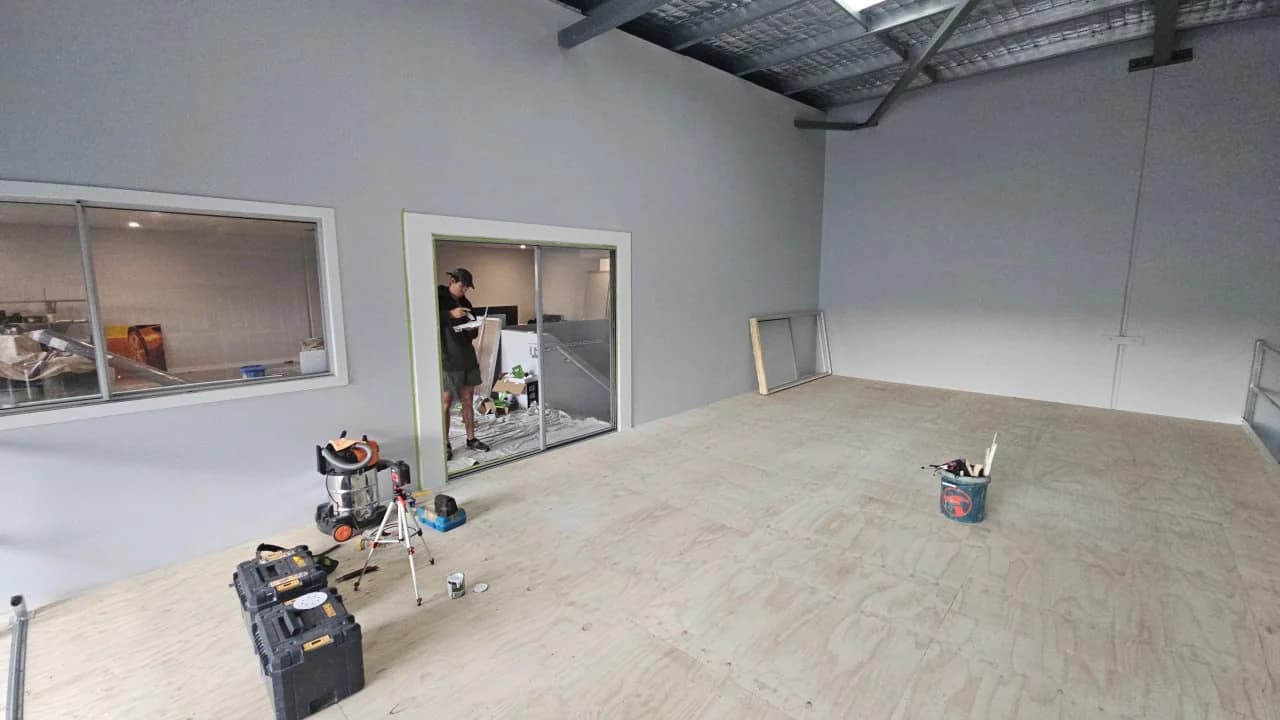 A warehouse fitout part way through, a glazed office and partition framed up on a plywood deck inside an industrial unit, with an internal window through to the space beyond it and tools and a vacuum on the floor