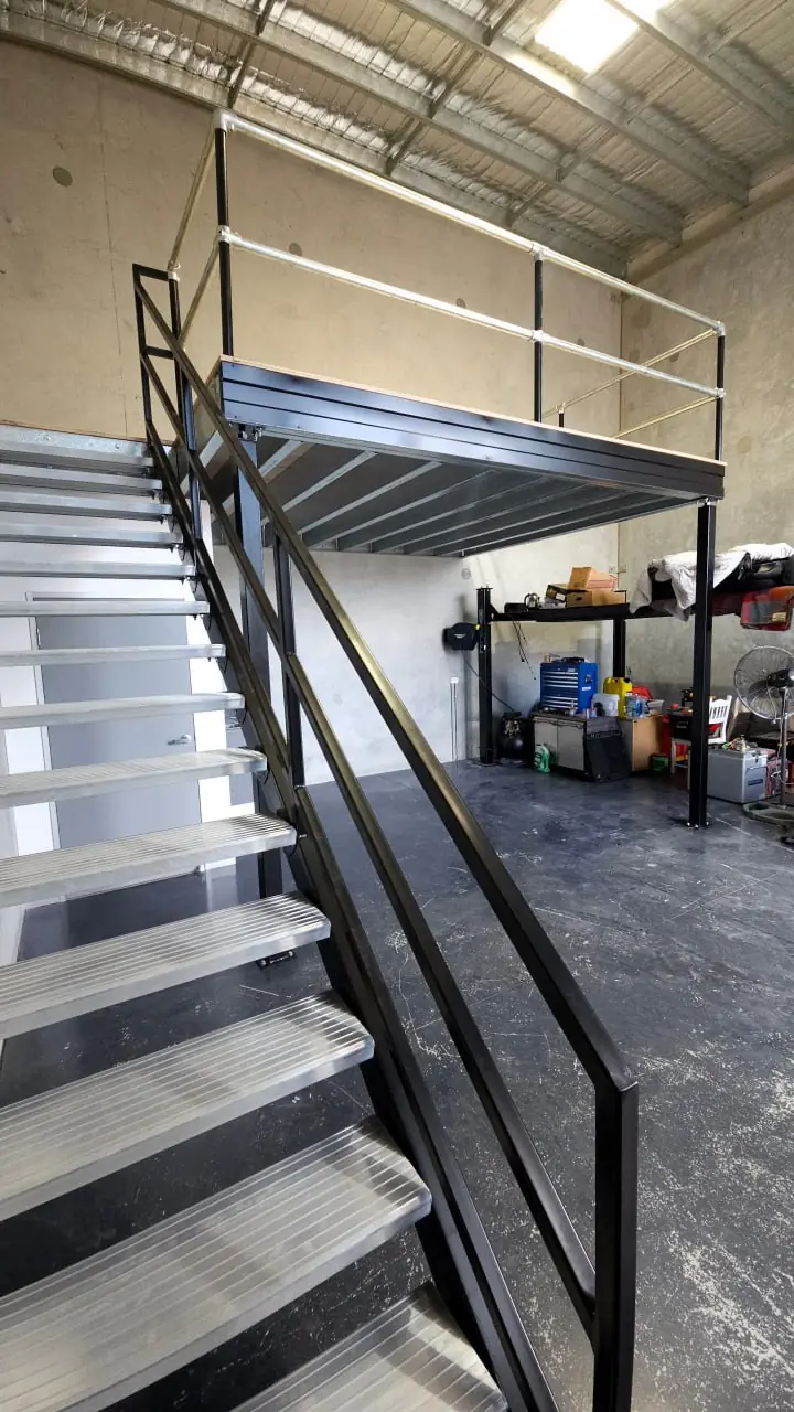 A black steel access stair with aluminium treads rising from a workshop floor to a mezzanine deck with a black edge beam, plywood decking and galvanised guardrail above