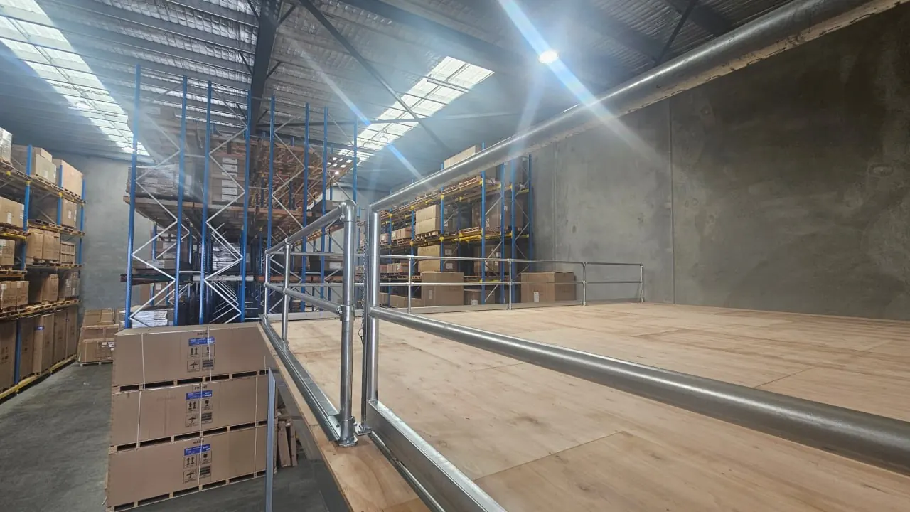 Looking along a finished mezzanine deck with a galvanised guardrail on its open edge, with loaded pallet racking and a warehouse roof beyond it