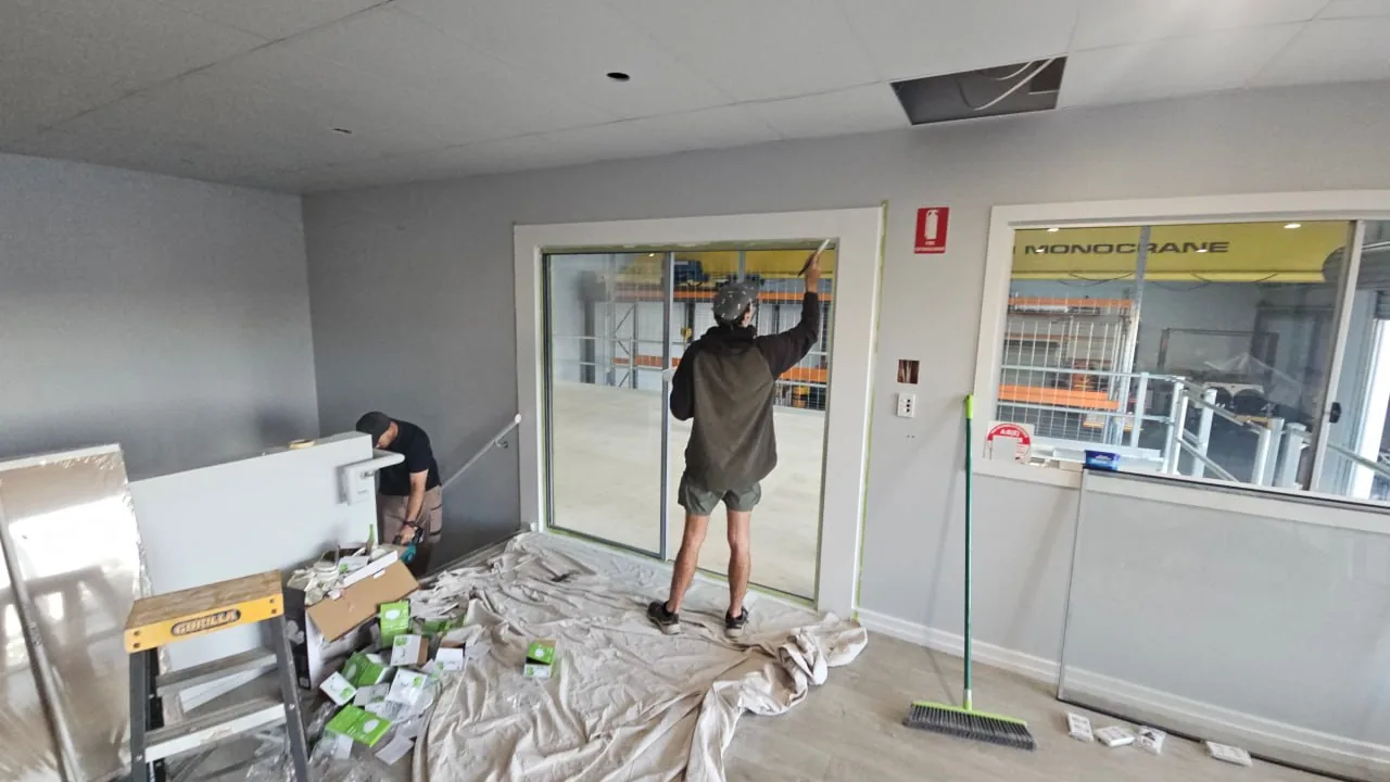 A mezzanine office being fitted out, with a carpenter working on the head of the glazed opening that looks down onto the warehouse floor and its overhead crane