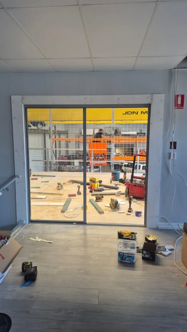 Inside a finished mezzanine office looking out through full height sliding glazing onto the warehouse floor below, with an overhead crane and orange pallet racking beyond the glass