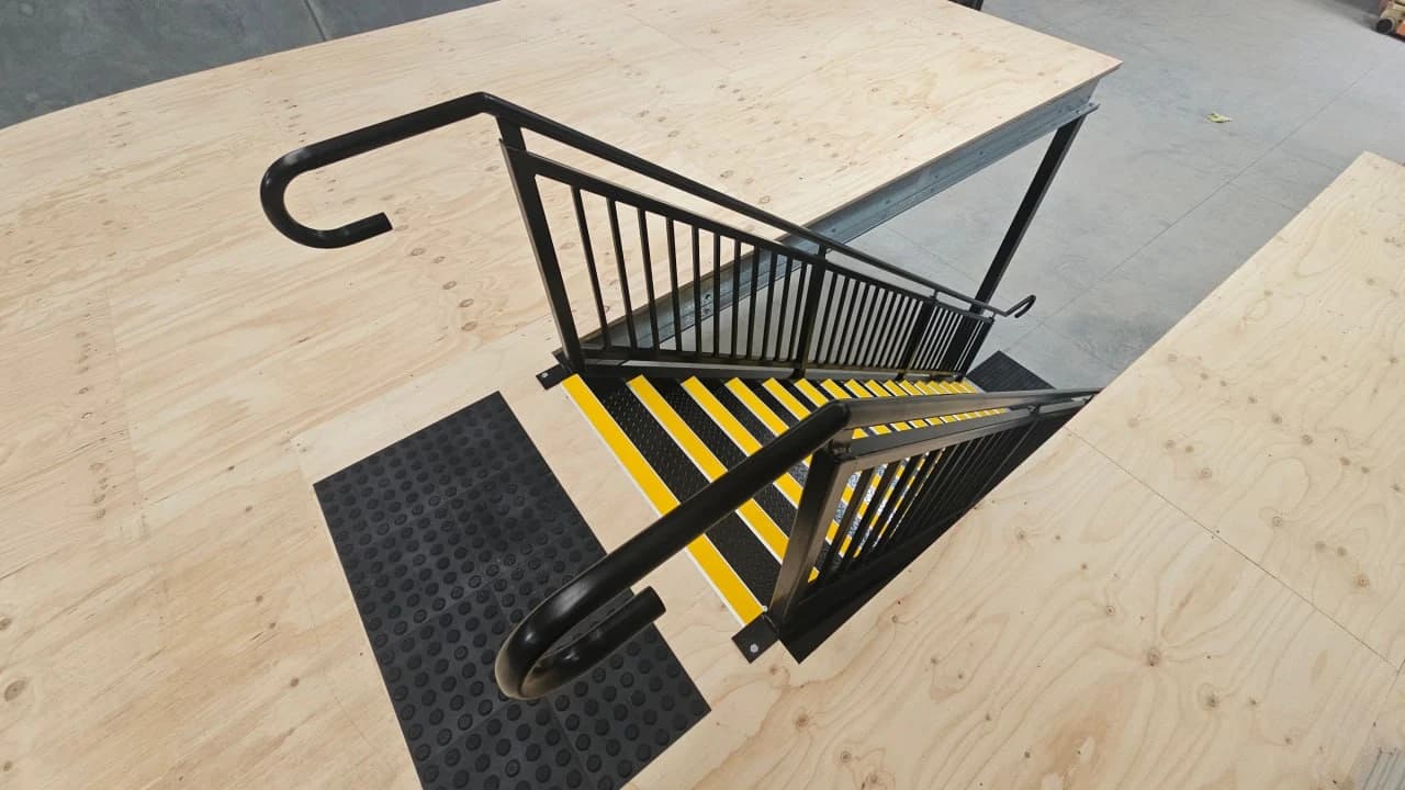 Looking down a black steel mezzanine stair from the deck, with yellow nosing on every checker plate tread, a returned handrail on both sides and a plywood mezzanine deck at the top