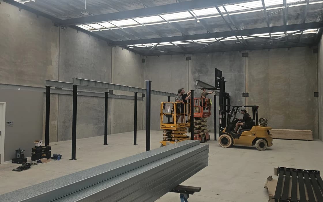 A mezzanine steel frame going up inside a bare industrial tenancy, black columns on base plates carrying galvanised beams, with two workers on a scissor lift and a forklift lifting the next beam into place