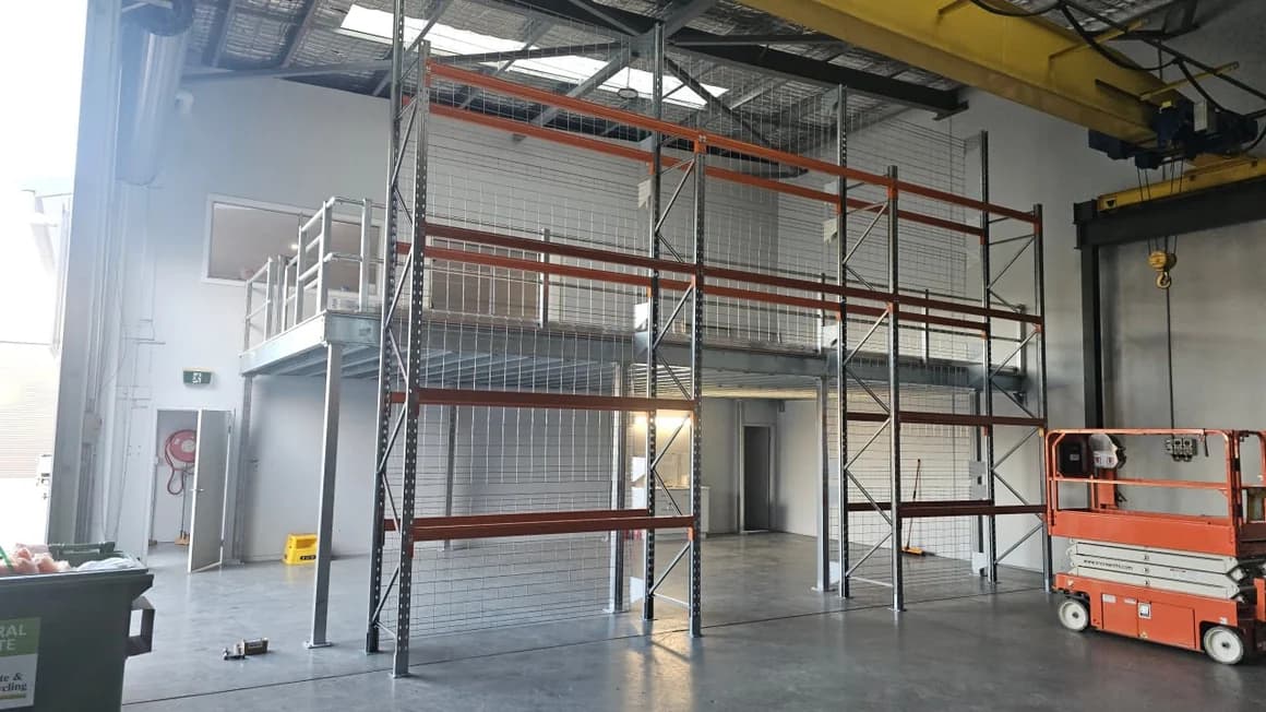 A steel mezzanine platform with a guardrail and access stair beside a tall run of pallet racking with orange beams and mesh infill, inside a warehouse with a gantry crane rail overhead
