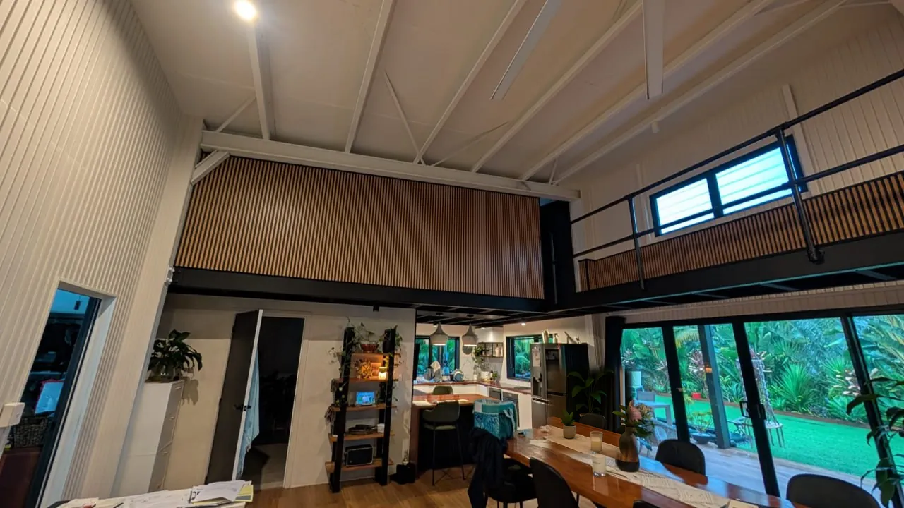 A residential mezzanine level under a raked ceiling, with a timber batten front on one side, a black steel edge beam beneath it, and a black steel balustrade with timber batten infill running along the open side, above an open plan kitchen and dining room that looks out to the garden