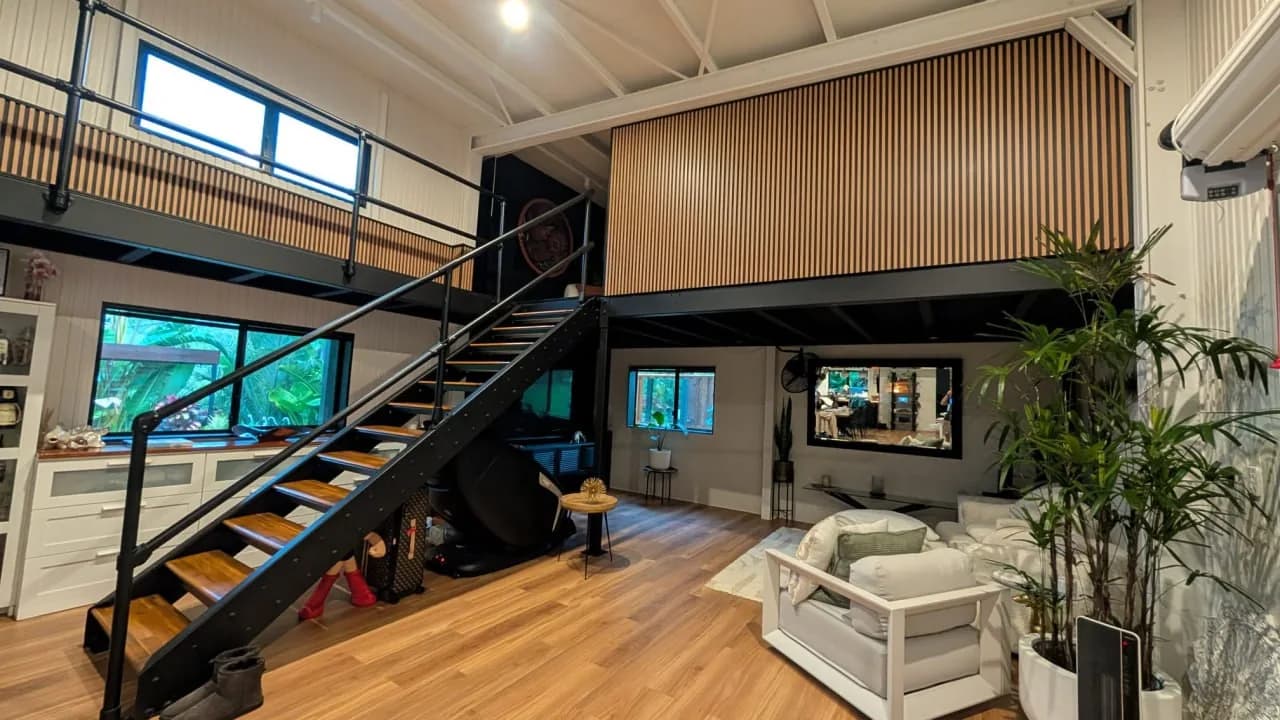 A black steel stair with timber treads rising to a mezzanine level inside a double height home, with a timber batten front above and an open plan living room below