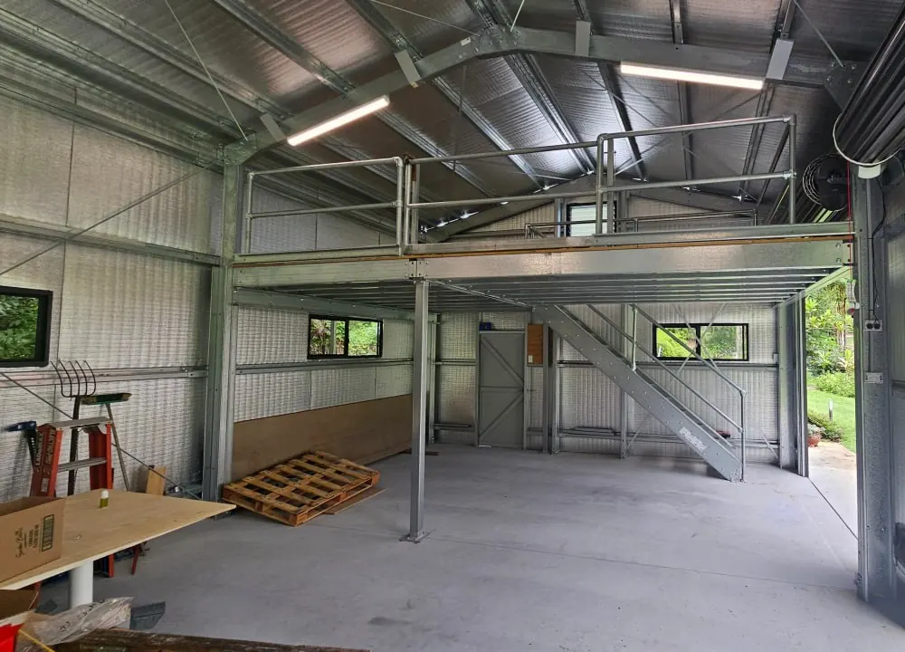 A galvanised steel mezzanine standing on its own posts inside a shed, with a tube guardrail along the deck edge, a straight run of stairs down to the concrete floor and the roller door open beside it