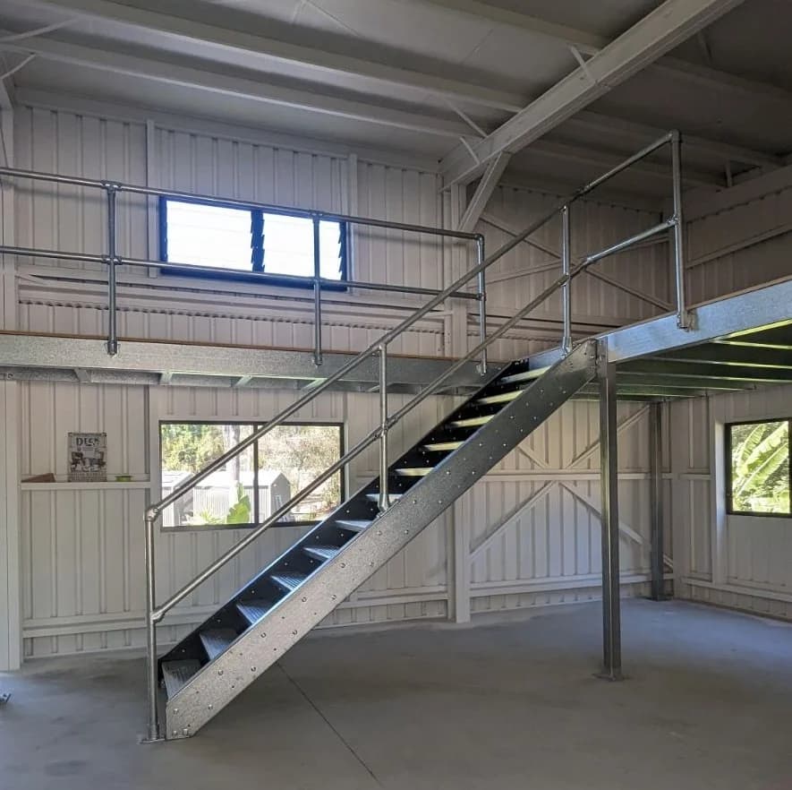 A galvanised steel mezzanine built inside a lined shed, with a tube guardrail along the deck edge and a straight run of checker plate stairs down to the concrete floor