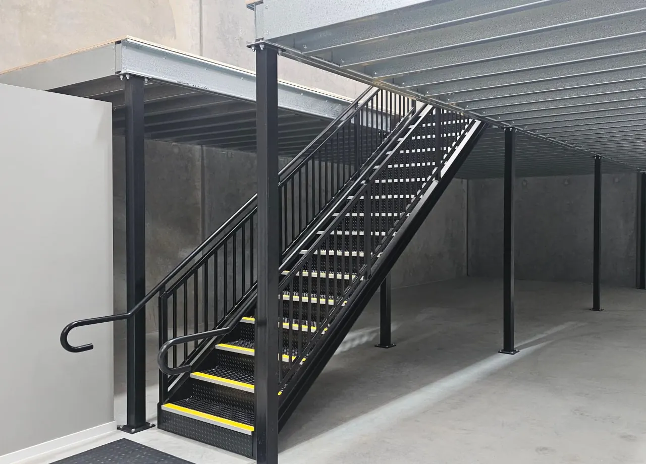 A black steel mezzanine stair with checker plate treads and yellow nosings, a handrail on both sides and a returned end, rising to a galvanised mezzanine deck in a Gold Coast warehouse