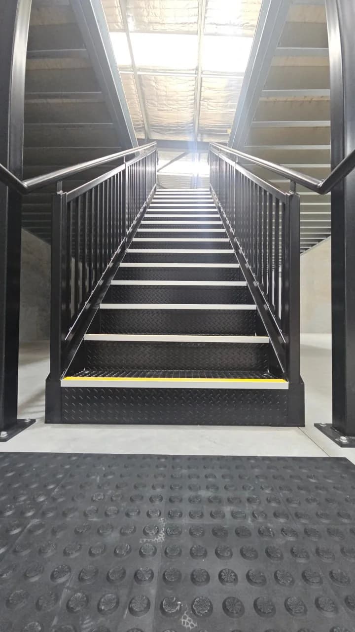 Looking straight up a black steel mezzanine stair with checker plate treads, yellow nosing on the bottom step and tactile ground indicators set into the floor at its base