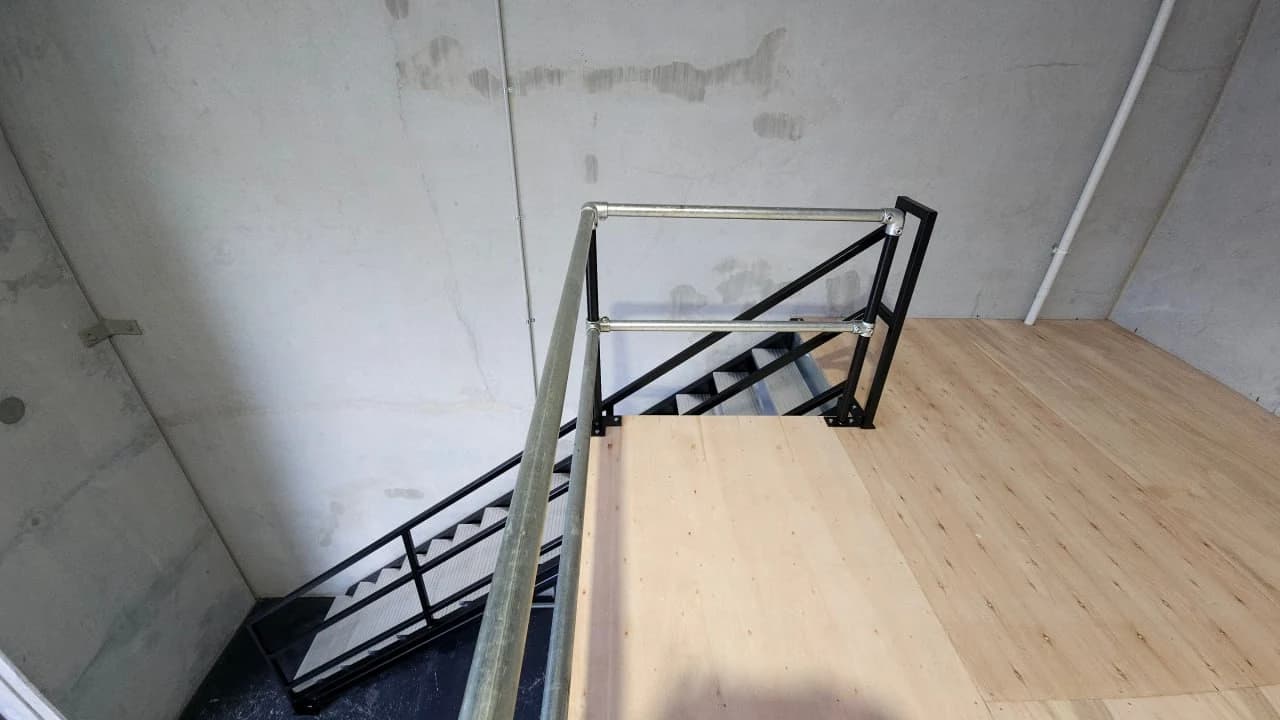 Looking down a new black steel access stair from the mezzanine deck it lands on, with a galvanised handrail turning across the landing and fresh plywood decking underfoot