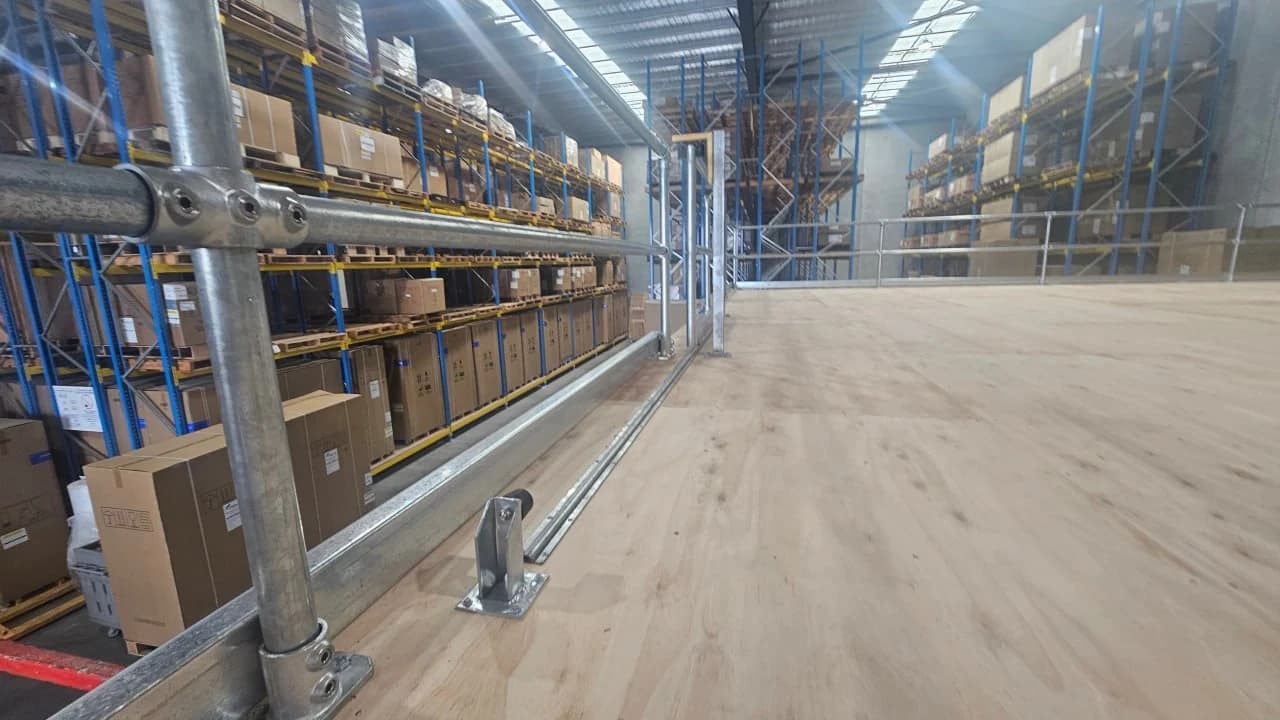 Looking along a finished mezzanine deck of plywood sheeting with a galvanised tube guardrail down one side, loaded pallet racking running beside it