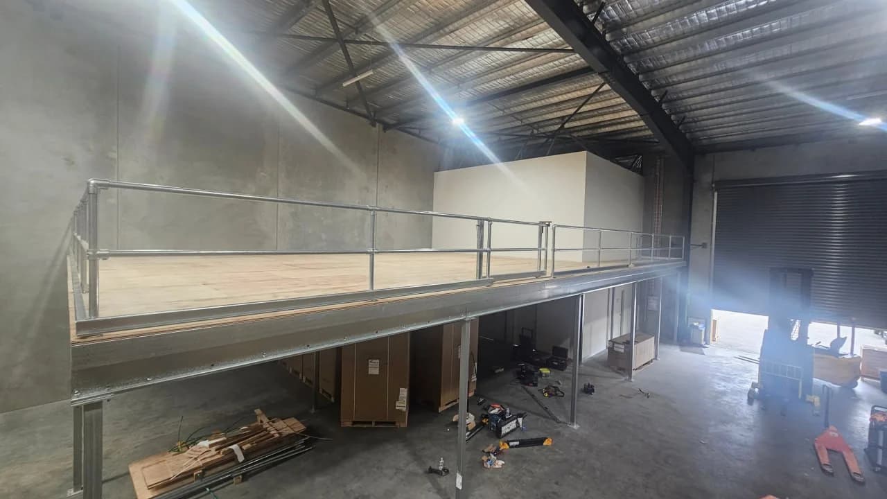 A completed warehouse mezzanine on steel columns with a galvanised edge beam and guardrail, the plywood deck above and pallets stored in the clear space underneath