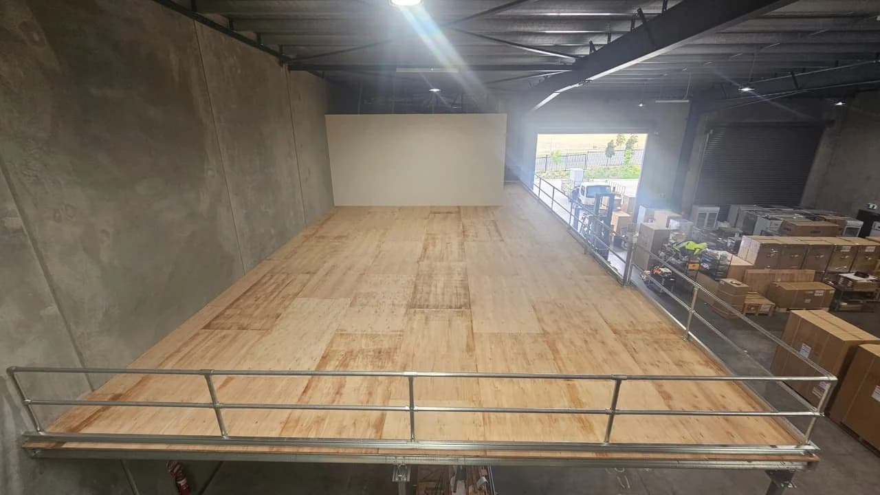 A completed Gold Coast mezzanine floor with a plywood deck and galvanised guardrail, looking along it from one end, with stock and a forklift on the warehouse floor below